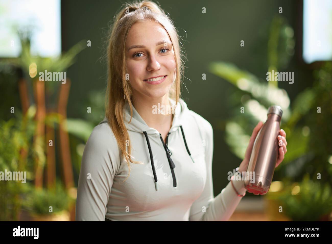Portrait of smiling modern woman in red fitness clothes with bottle of water in the modern green living room. Stock Photo