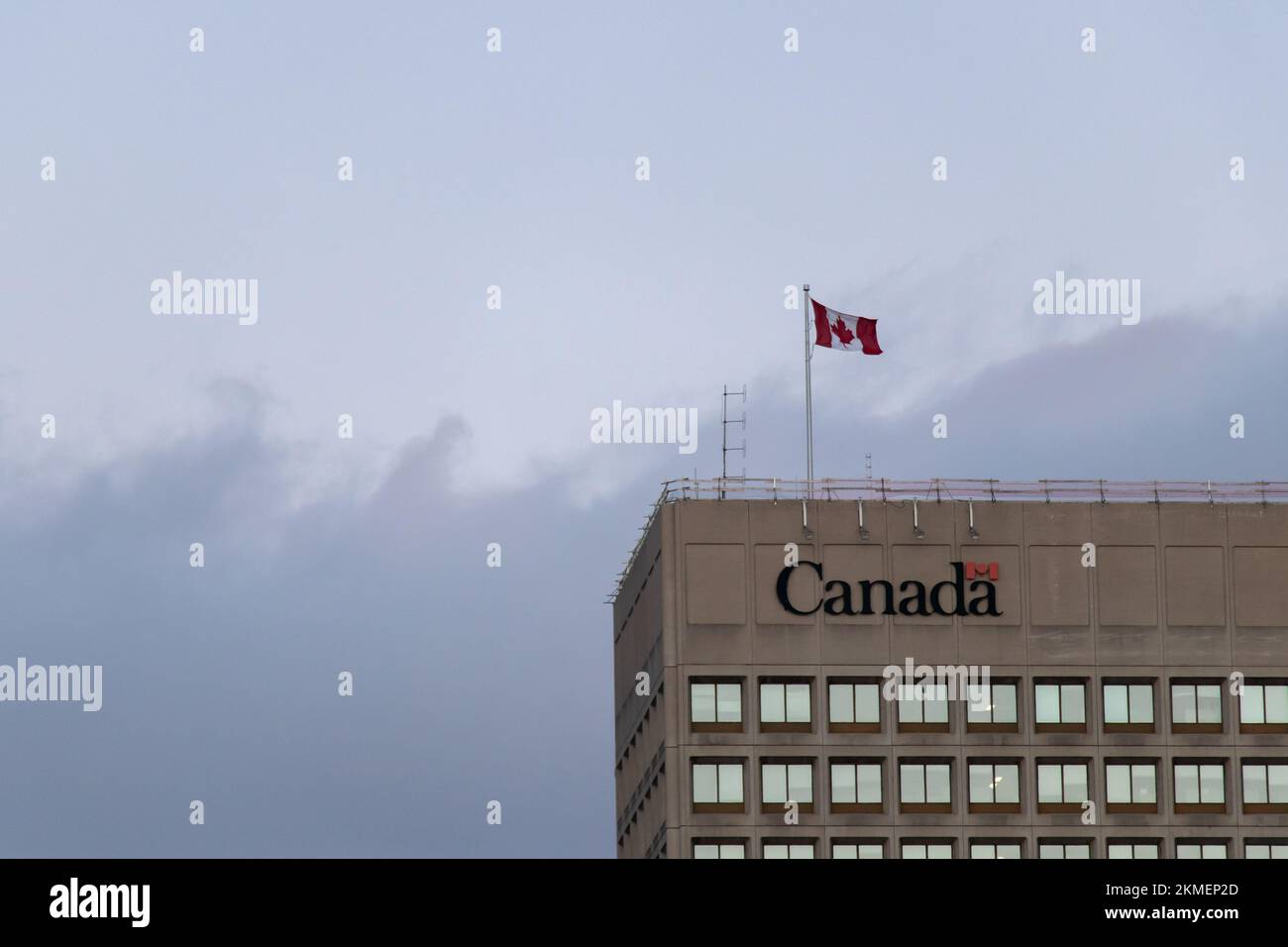The Government of Canada logo is atop of a tall office building in downtown Ottawa, while the Canadian flag is seen flying above on windy afternoon. Stock Photo