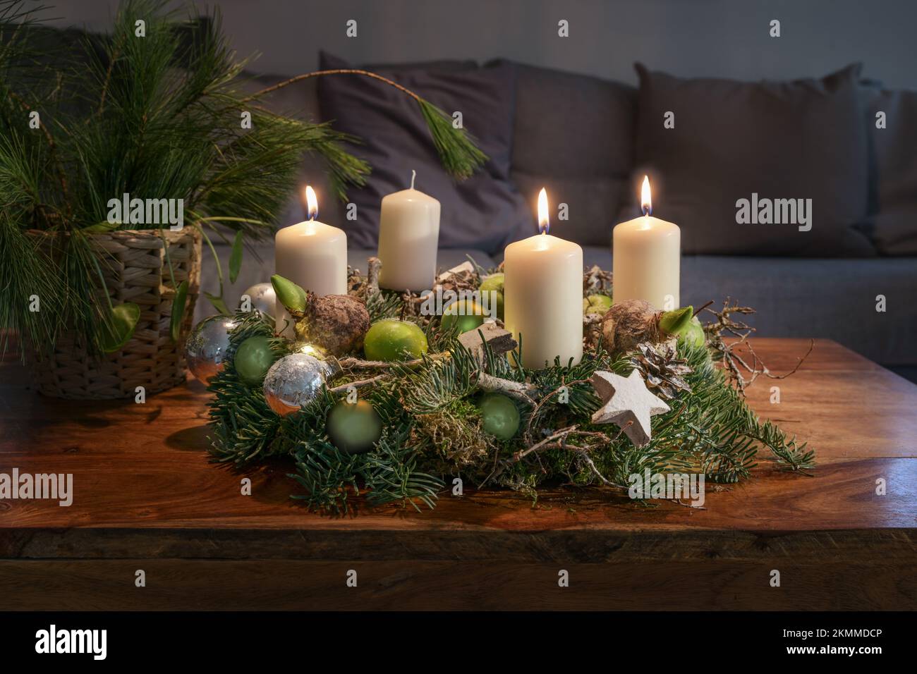 Third Advent, wreath with three burning white candles and Christmas decoration on a wooden table in front of the couch, festive home decor, copy space Stock Photo