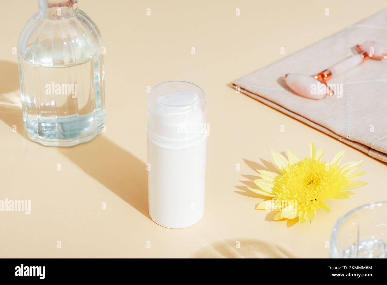 White cosmetic bottle, yellow flower and facial roller on beige background in sunlight. Skincare, spa and wellness concept. Selective focus. Stock Photo