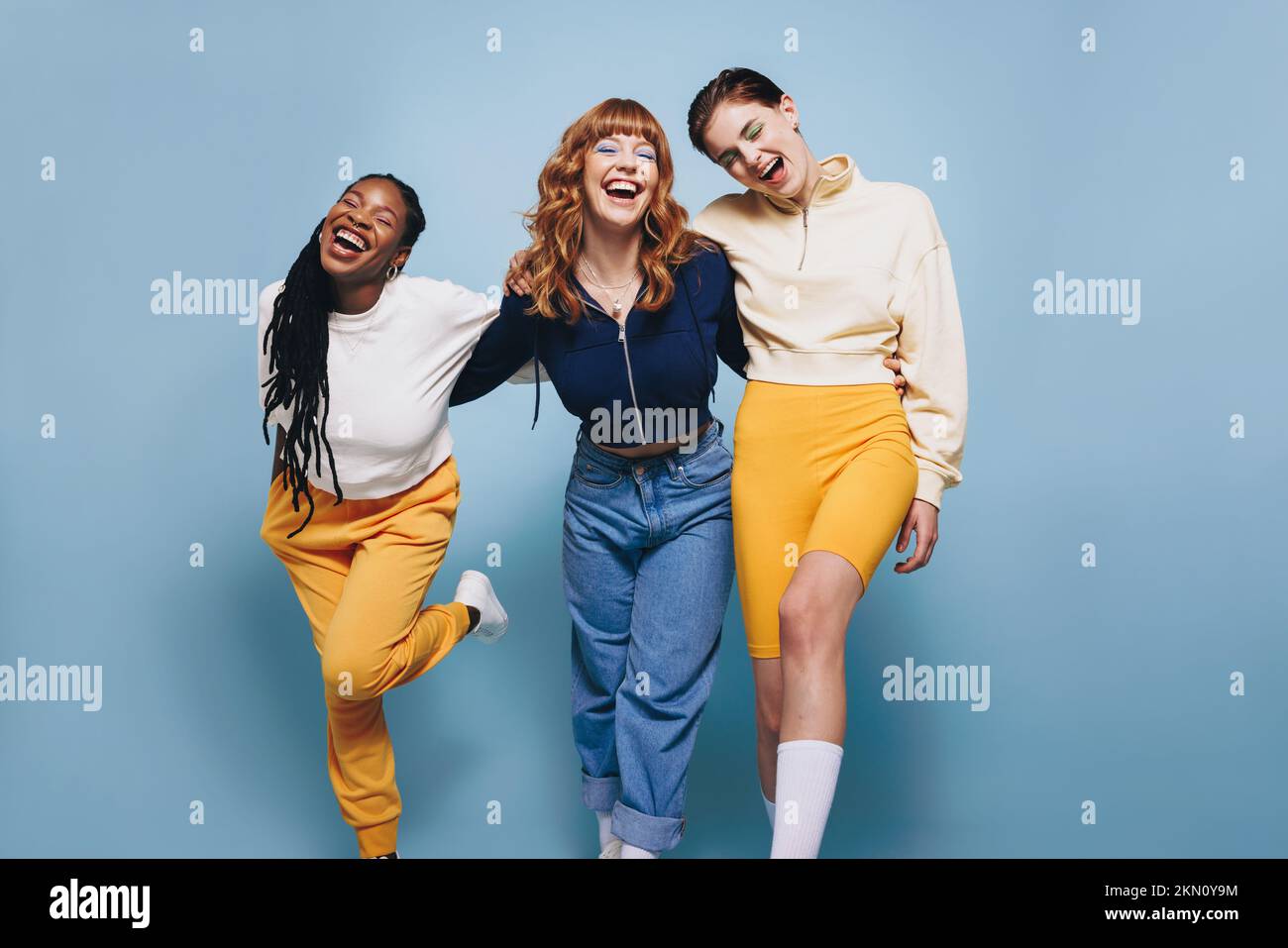 Three female friends laughing happily while embracing each other. Group of cheerful young women having fun while standing together against a blue stud Stock Photo