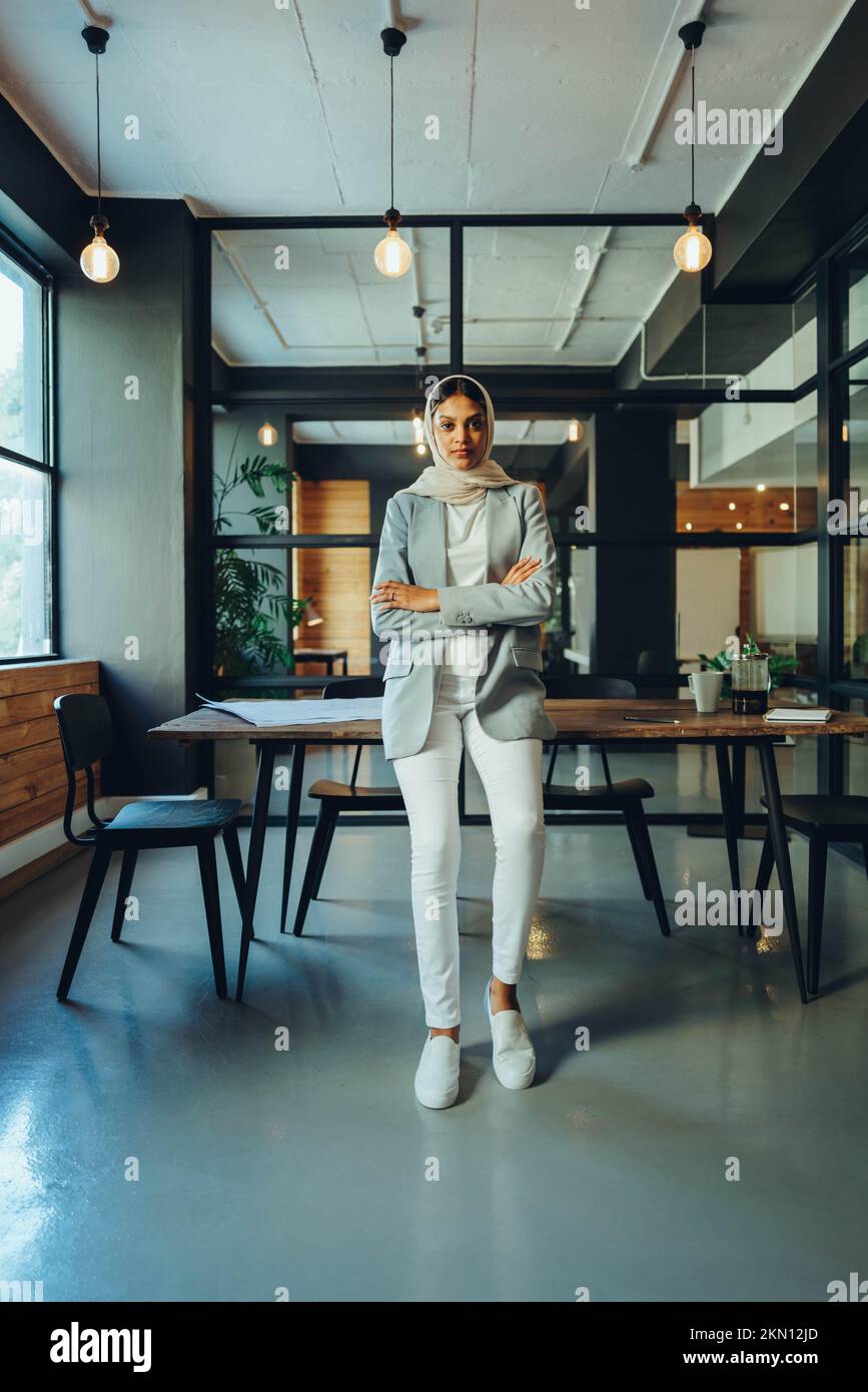 Young Muslim businesswoman looking at the camera while standing in an office boardroom. Self-confident female entrepreneur wearing a hijab in a modern Stock Photo