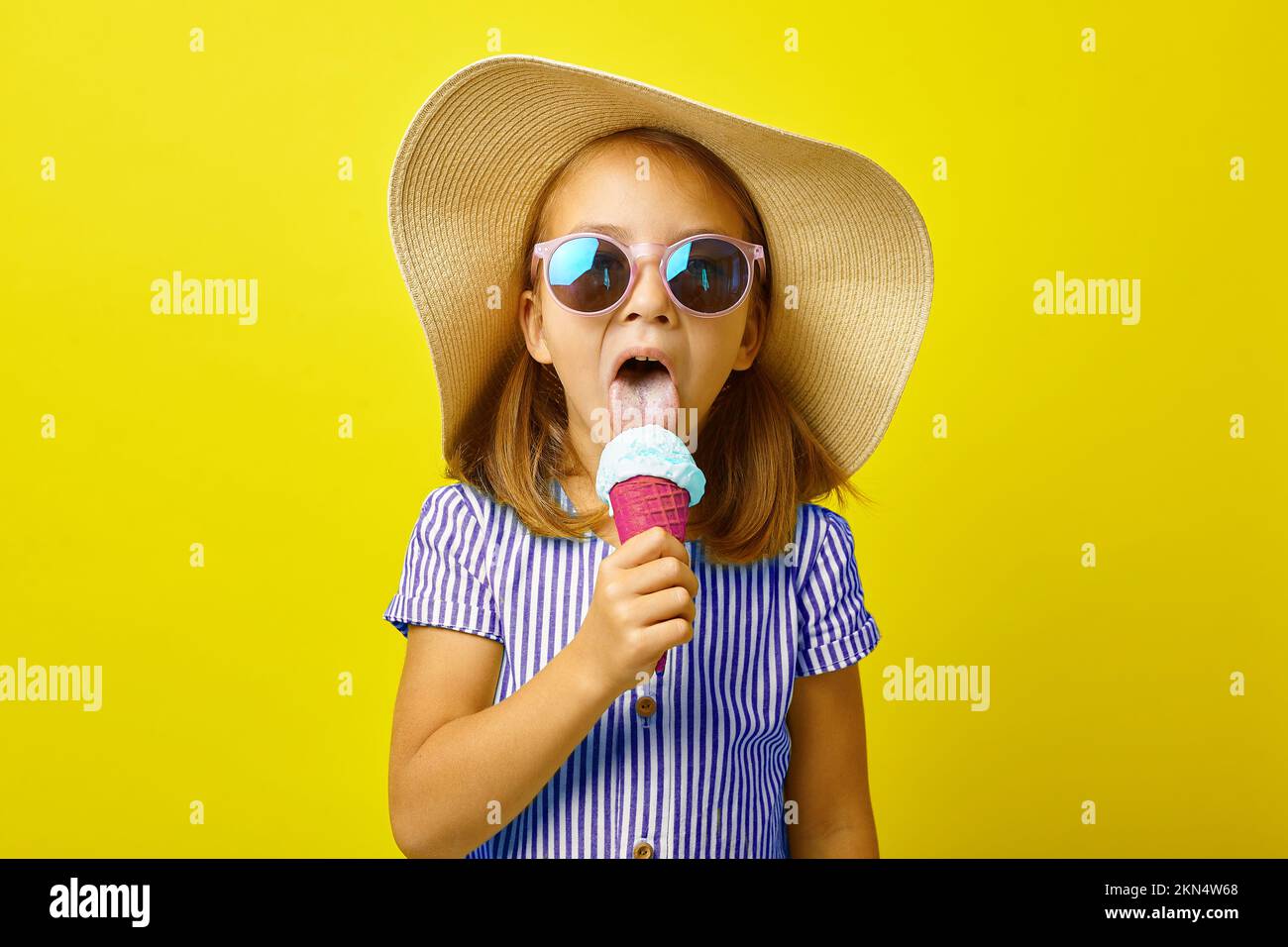 Funny little girl in summer clothes eating beautiful ice cream, standing on yellow isolated background. Stock Photo