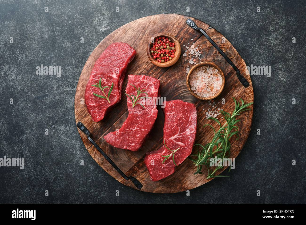 Raw steaks. Top blade steaks on wood burning board with spices, rosemary, vegetables and ingredients for cooking on black background. Top view. Copy s Stock Photo