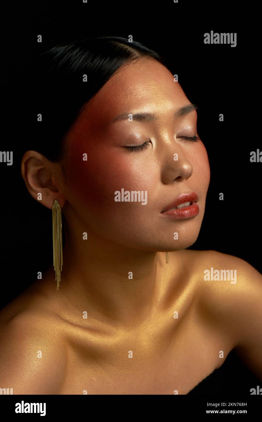 An Asian girl on a black background in the studio. Gold. Relaxed state. Self-care and enjoyment. Femininity. Stock Photo