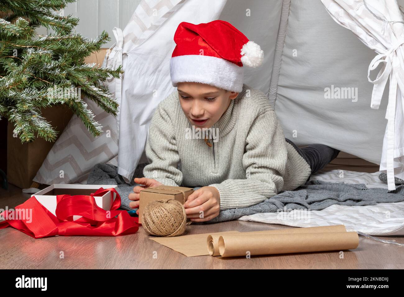 Family Christmas. Children open Christmas presents. A teenage boy in a Santa hat opens a gift and smiles while sitting on the floor of the house Stock Photo