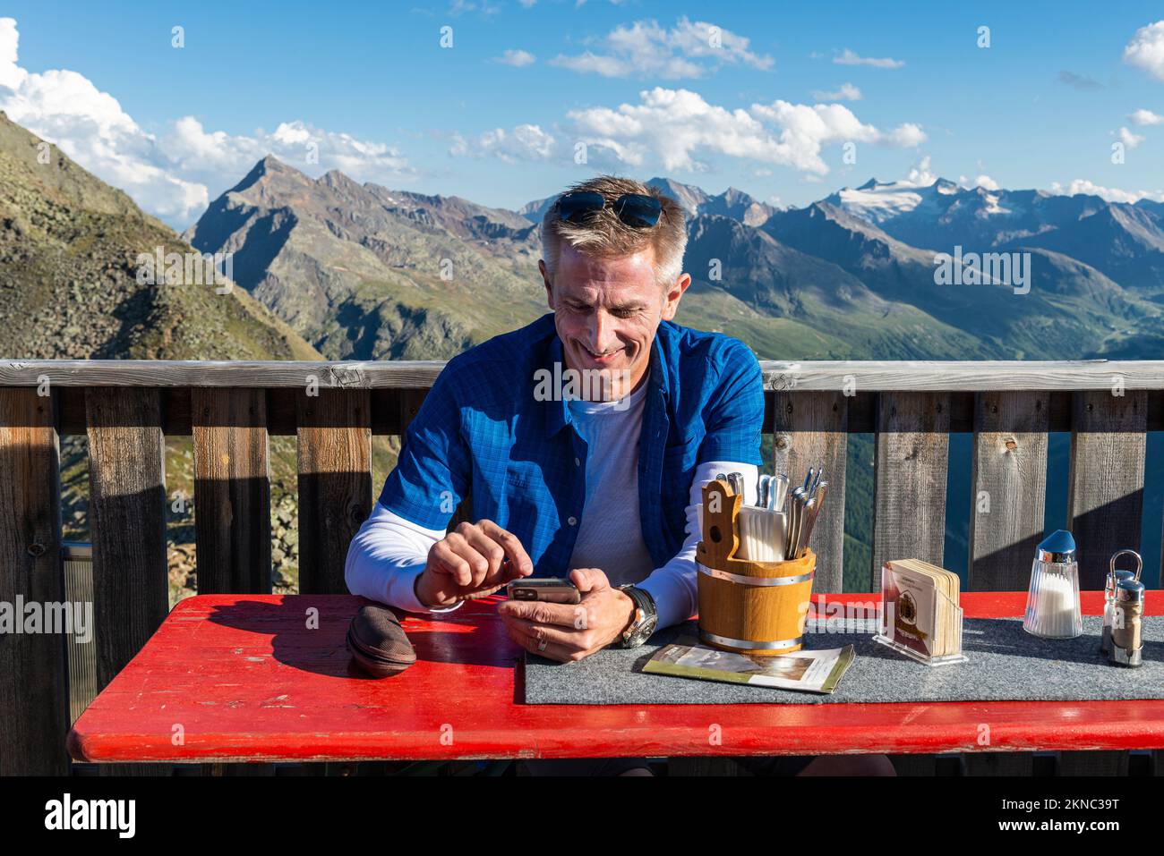 A hiker sits on the terrace at the Brunnenkogelhaus mountan hut and looks at his mobile phone in front of the Ötztal Alps and Gurgl Valley,Austria Stock Photo