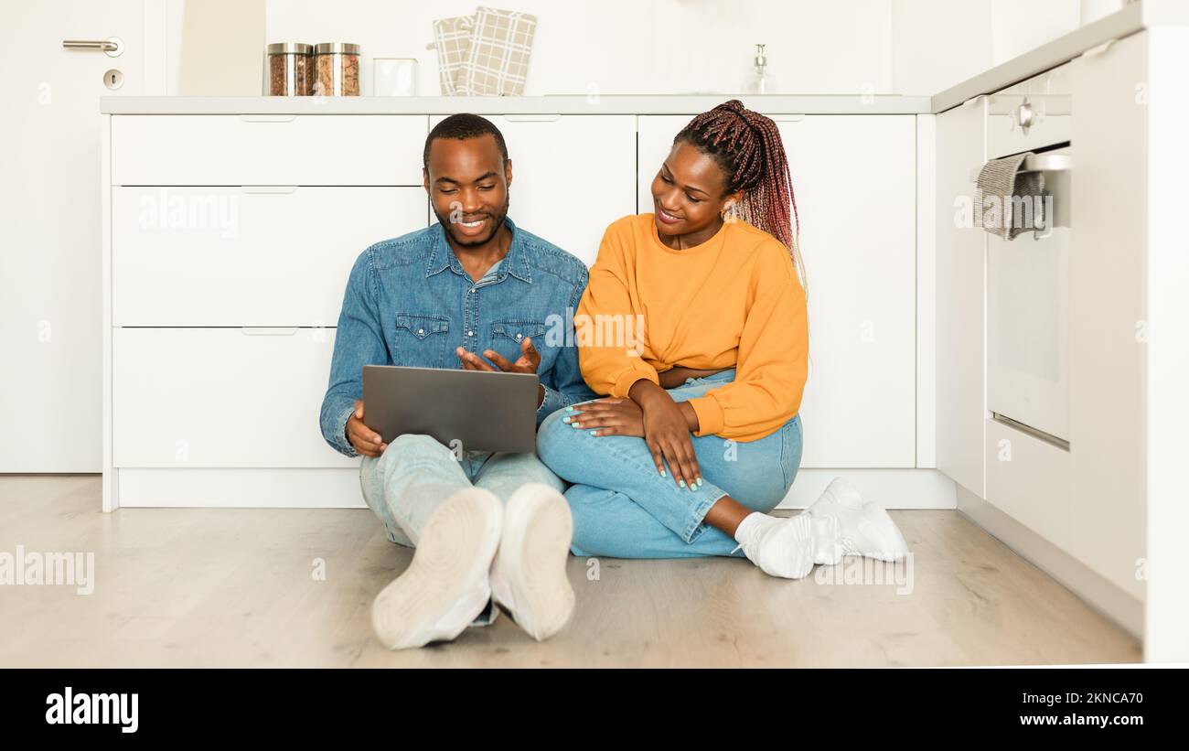 Excited african american spouses with laptop sitting on floor in kitchen, shopping online or browsing internet Stock Photo