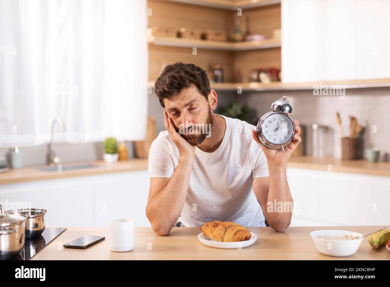 Sad tired middle aged caucasian male with beard in white t-shirt shows alarm clock and sleeps in modern kitchen Stock Photo