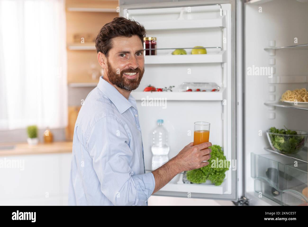 Happy handsome adult caucasian man with beard in shirt opens refrigerator door, takes glass of juice Stock Photo