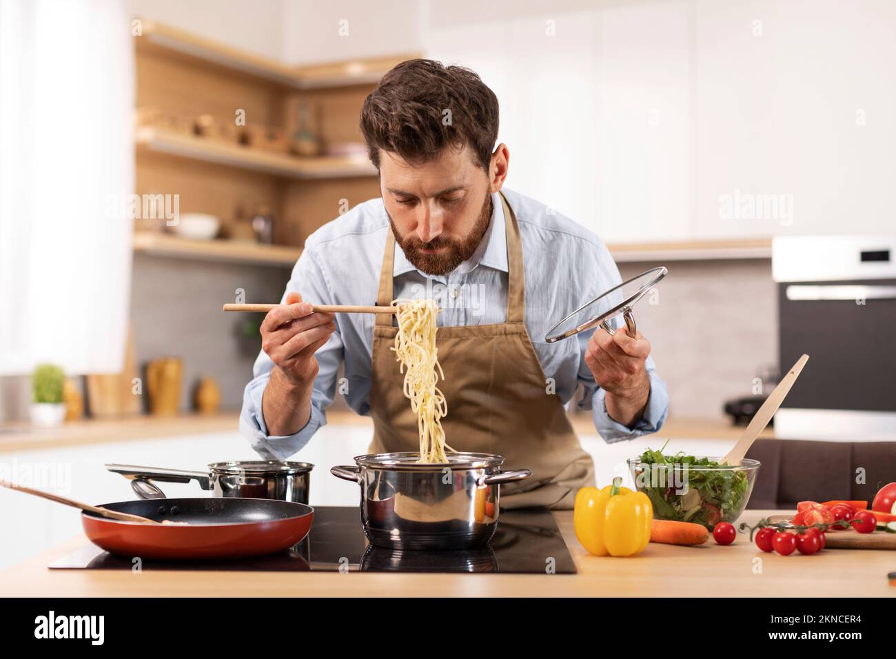 Serious handsome mature caucasian man with beard in apron prepare pasta in minimalist kitchen interior Stock Photo
