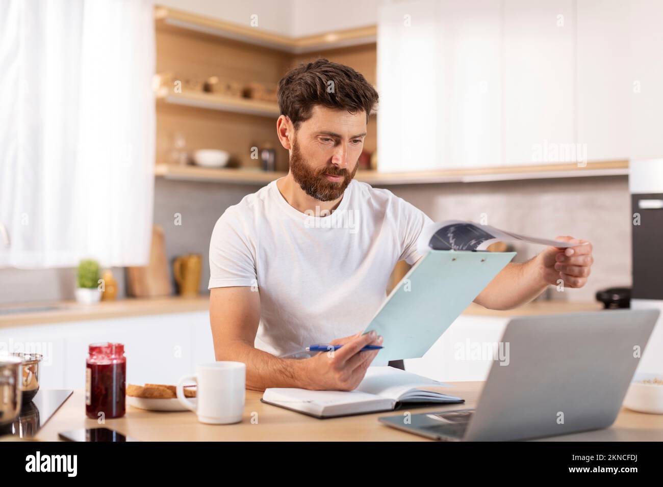 Serious adult caucasian male with beard works with documents and laptop in minimalist kitchen interior Stock Photo
