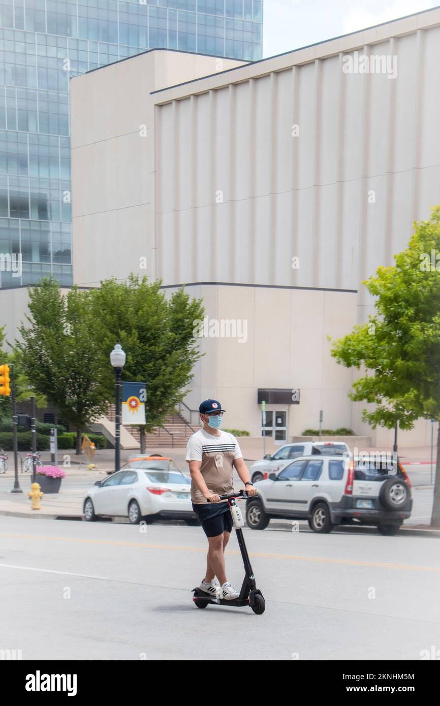 07 25 2020 Tulsa USA - The new normal - A young man in shorts and tee shirt and ball cap and face mask rides electric scooter down city street with ta Stock Photo