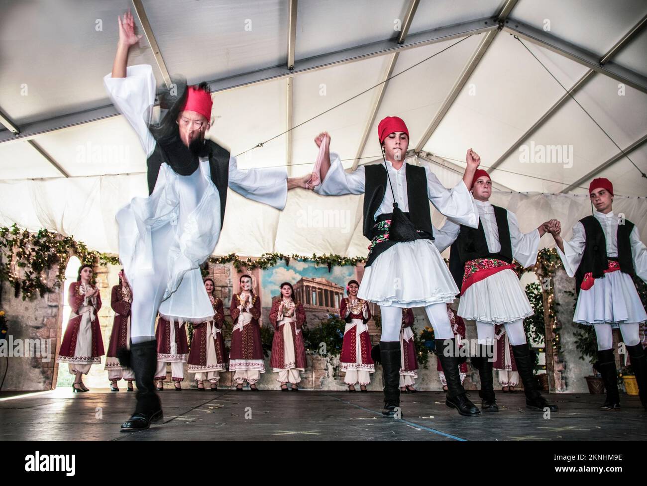 09-21-2019 Tulsa USA - Costumed male Greek dancers in decorated tent venue  featuring dramatic kick at Greek Festival in Tulsa with women dancers clap Stock Photo