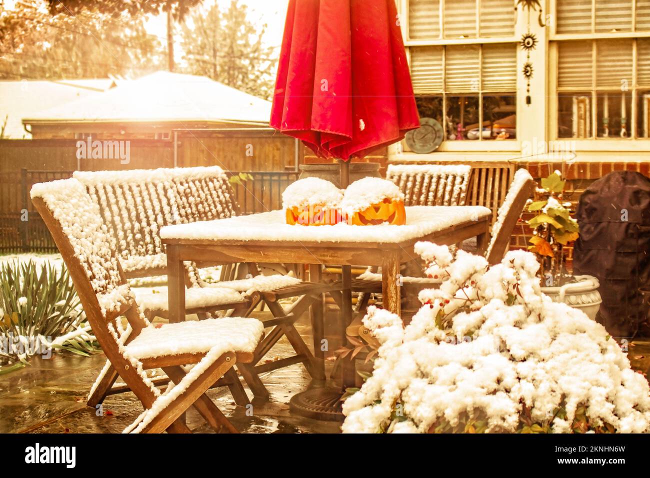 Early snow - outdoor table and chairs with two jack o lanterns and a sun umbrella on a patio during a snow shower with house windows and urban neighbo Stock Photo