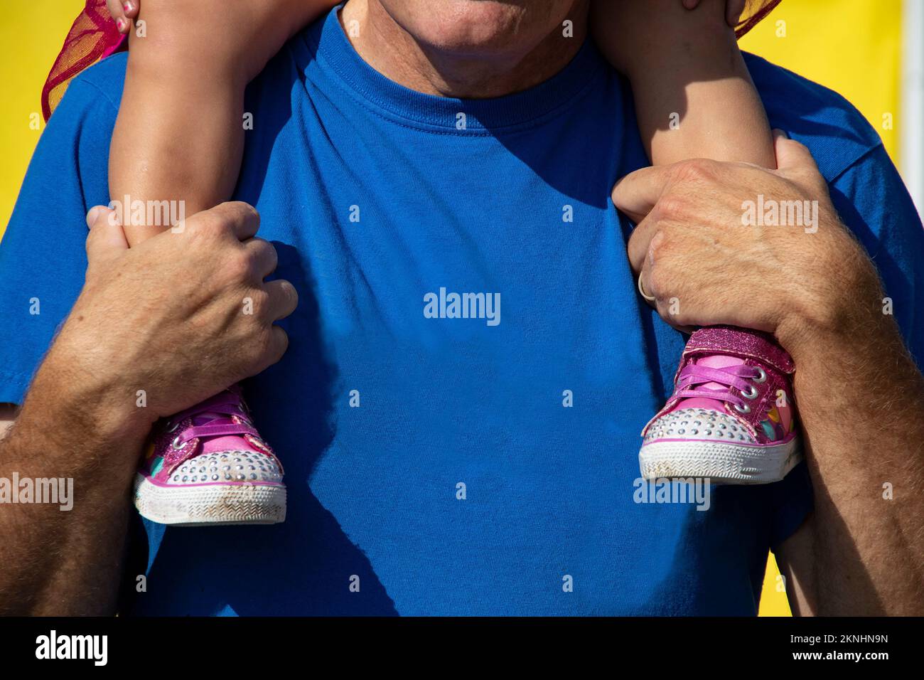 Father or grandfathe in blue teeshirt with little girl with pink sneakers on his shoulders - showing hands holding her legs- unrecognizable Stock Photo