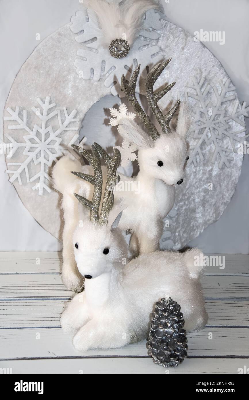 Sparkly fluffy white Christmas snow deer decorations in front of white wreath on white planks with silver pine cone - Selective focus Stock Photo