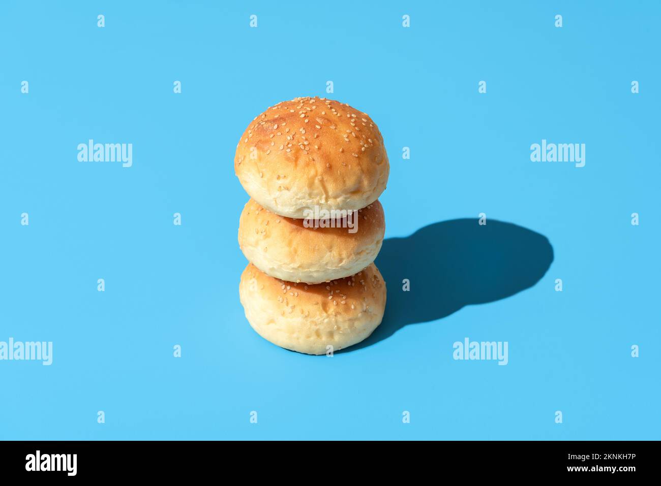 Stack of bread buns on a blue-colored table. Homemade burger buns with sesame seeds minimalist on a colorful background Stock Photo