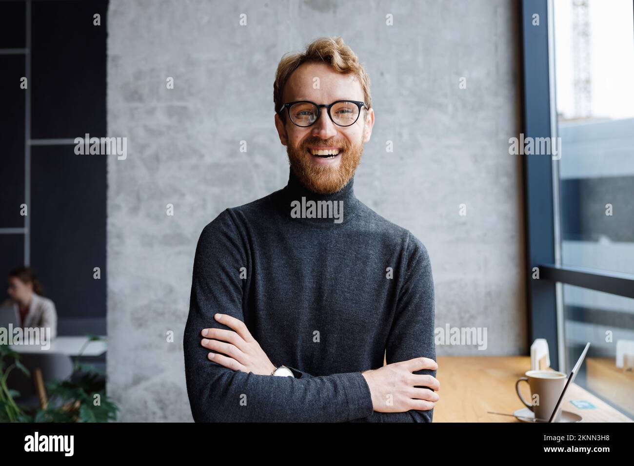 A young red-haired guy, a programmer or an entrepreneur in glasses, in a stylish cafe Looks at the camera and smiles. Freelancer works remotely Stock Photo
