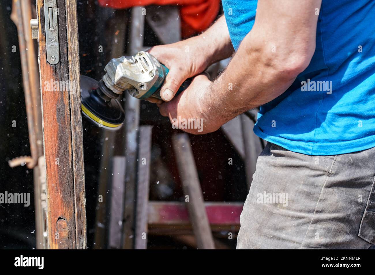 Man polishing wooden door with old angle grinder during sunny day, closeup detail to hands without gloves, fine wood dust flying in air Stock Photo
