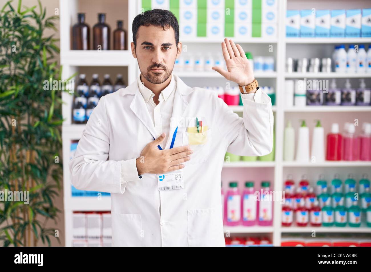 Handsome hispanic man working at pharmacy drugstore swearing with hand on chest and open palm, making a loyalty promise oath Stock Photo