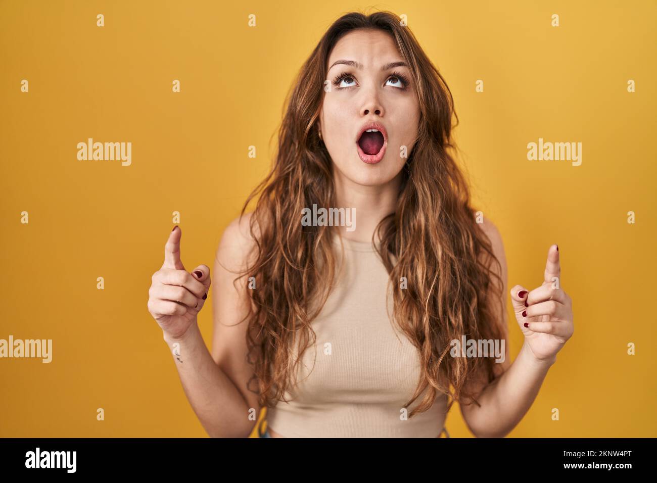 Young hispanic woman standing over yellow background amazed and ...