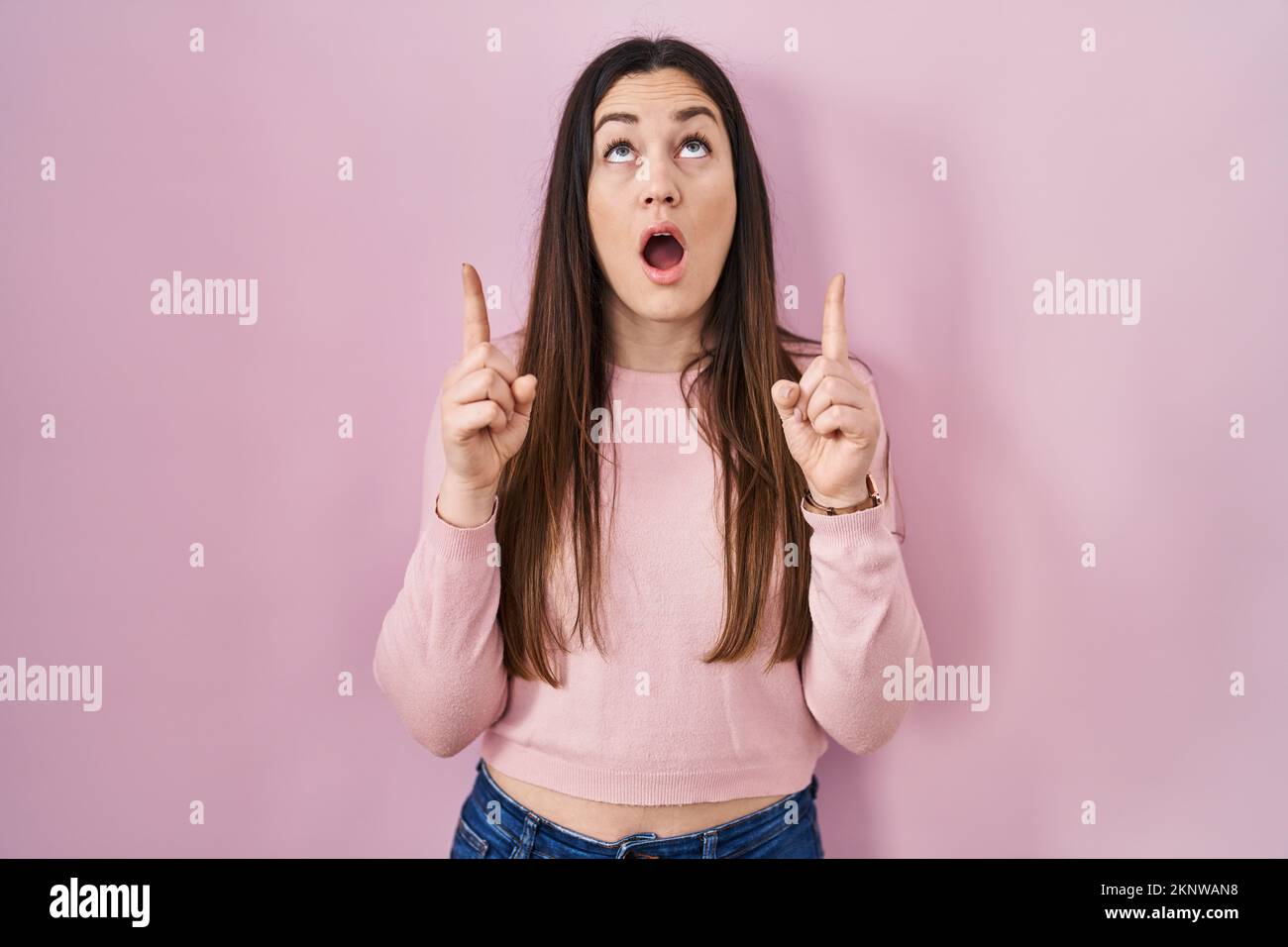 Young brunette woman standing over pink background amazed and surprised ...