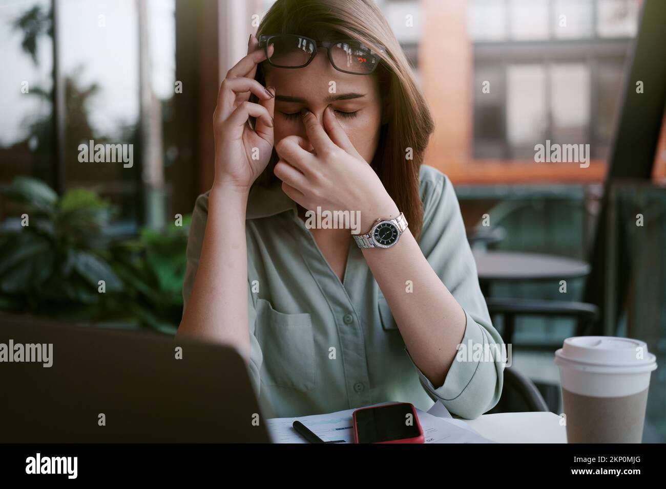 A tired and stressed young woman holds glasses and massages his nose while working hard in a cafe. Stock Photo