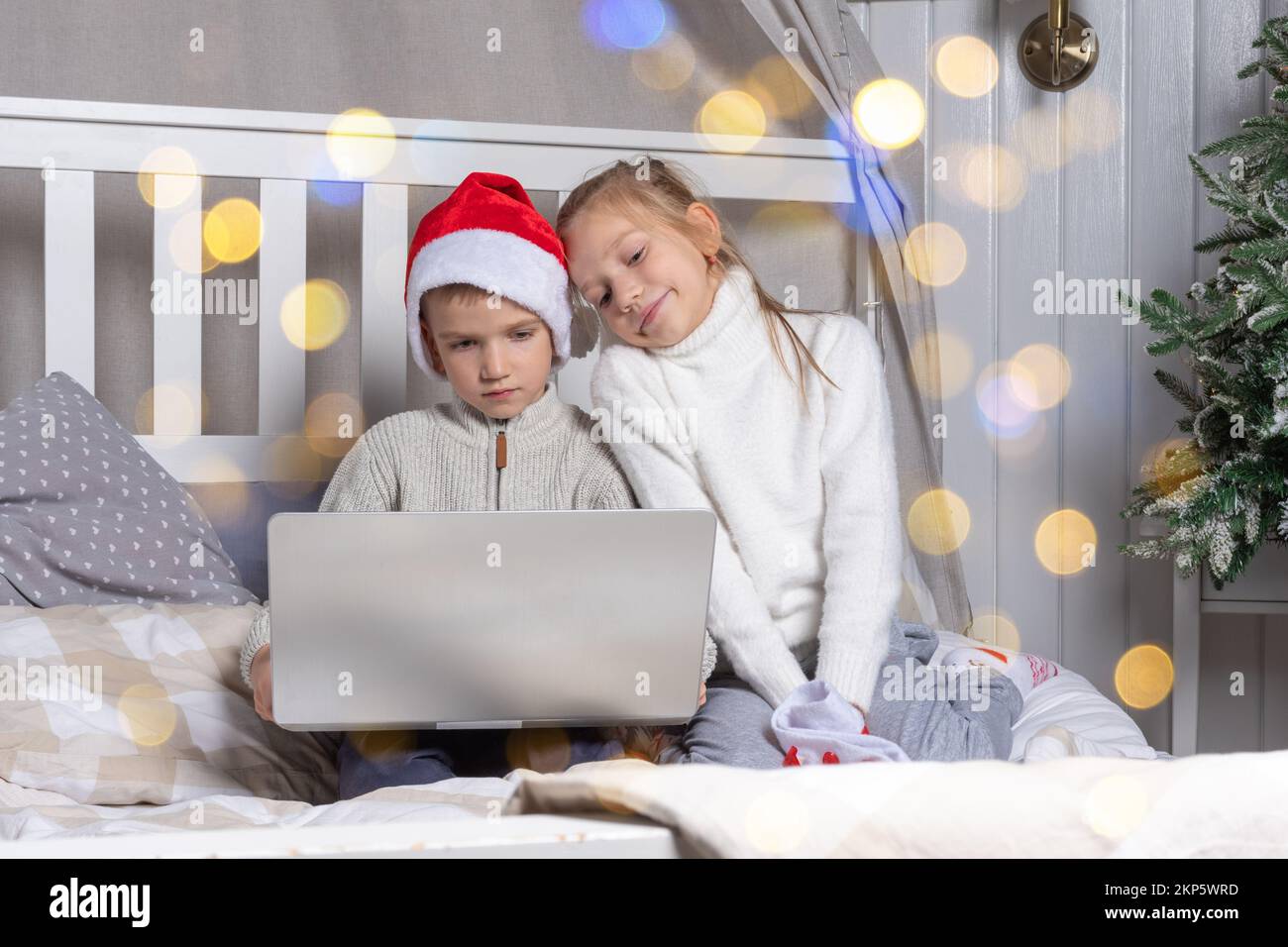 Happy kids in red Santa hats with a laptop on Christmas Eve at home. A boy and a girl online congratulate relatives sitting on the bed. Family x-mas e Stock Photo