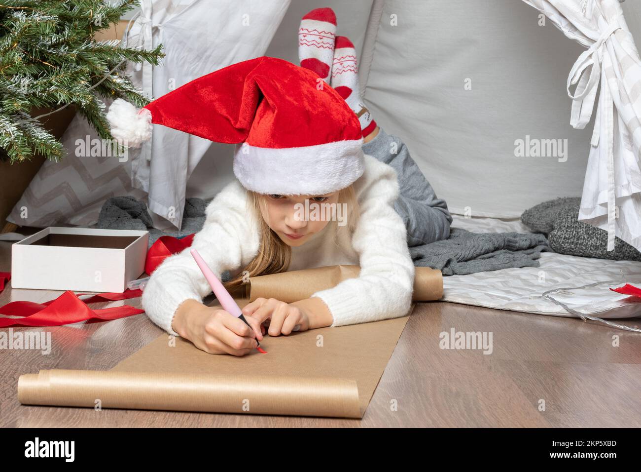 Christmas miracle wish list. Smiling girl in Santa Claus hat and a white sweater writing letter dreams for gifts to Santa Claus. The child hopes for a Stock Photo