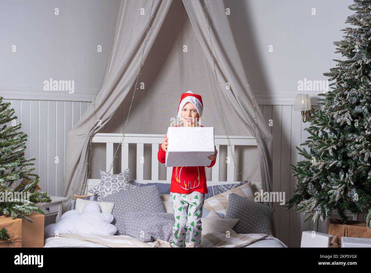 Happy funny child girl in pajamas and Santa hat holds a gift box and jumps in bed on Christmas morning. Children are enjoying the Christmas holiday. T Stock Photo