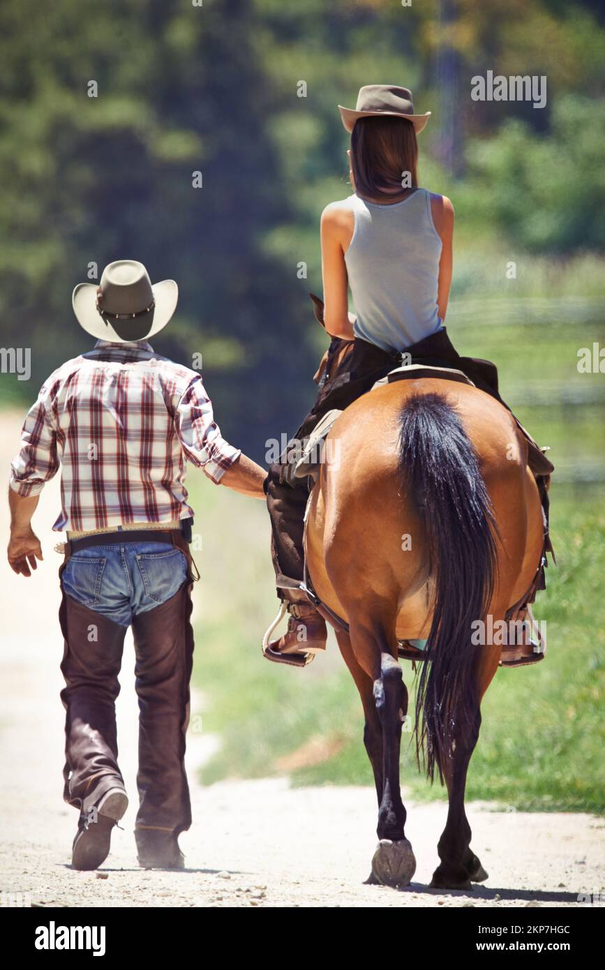 Loving the outdoors. Rear view of a cowboy leading a young woman on a horse along a country lane. Stock Photo
