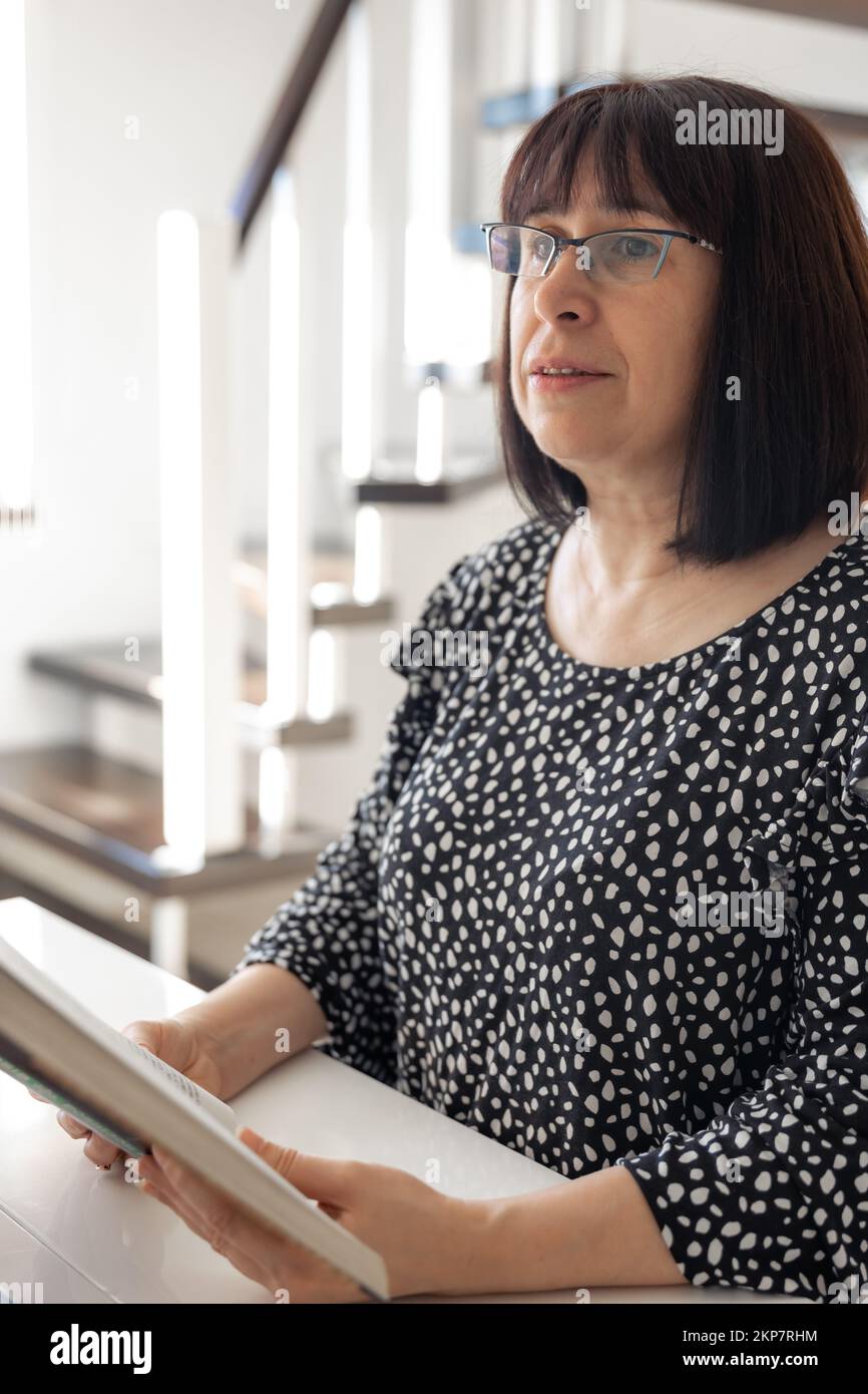 A middle-aged brunette woman in glasses with a bob haircut is reading a book. Stock Photo