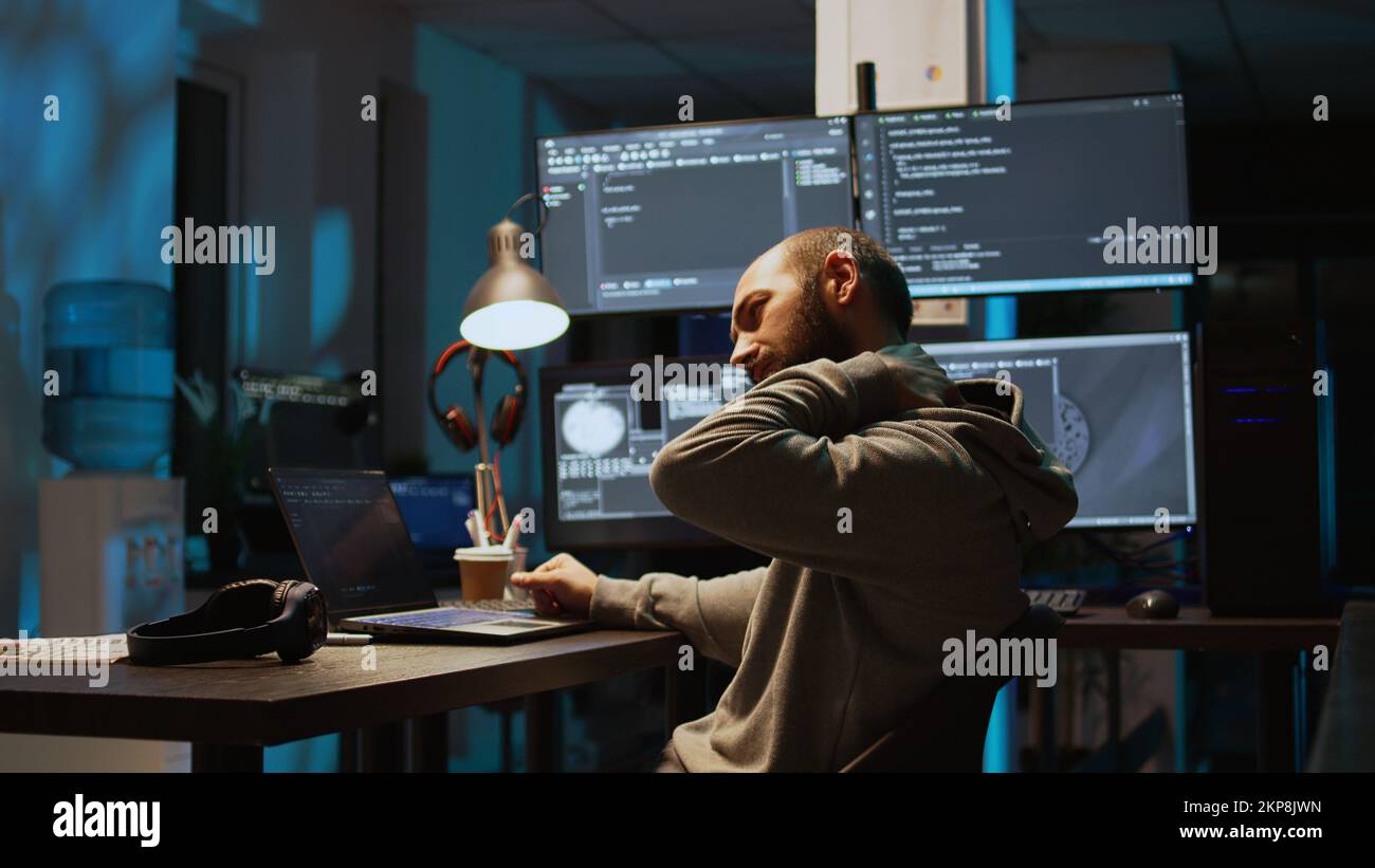 Male web programer writing code and dealing with back pain, sitting at office and feeling spinal discomfort at night. App developer coding html script on terminal window. Handheld shot. Stock Photo