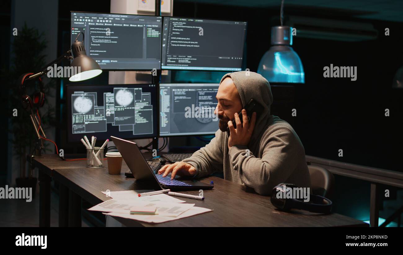 Young programmer talking on phone call in IT developing office, using smartphone for remote conversation. Male engineer working on html code developing, holding mobile phone. Handheld shot. Stock Photo