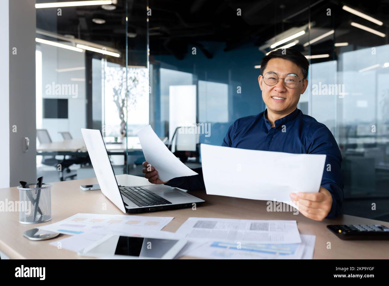Portrait successful satisfied asian financier accountant, man in shirt and glasses working inside office using calculator and laptop for accounts and financial documents reports, looking at camera. Stock Photo