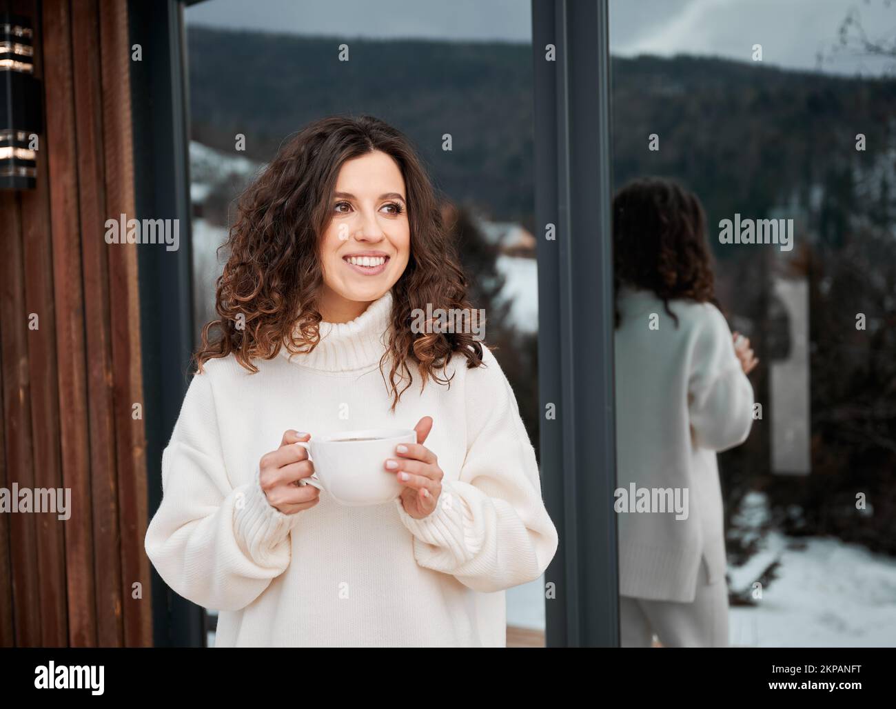 Curly young woman resting on terrace of modern barn house in the mountains. Happy female tourist holding cup of tea, enjoying in new cottage in winter. Stock Photo