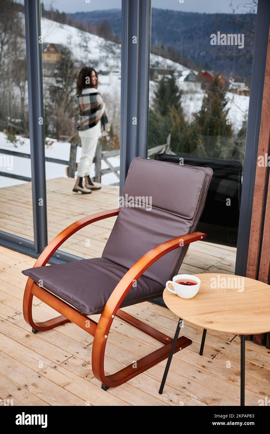 Chair and table with cup of tea on on terrace of modern barn house in winter mountains. Happy woman tourist reflected in huge window. Stock Photo