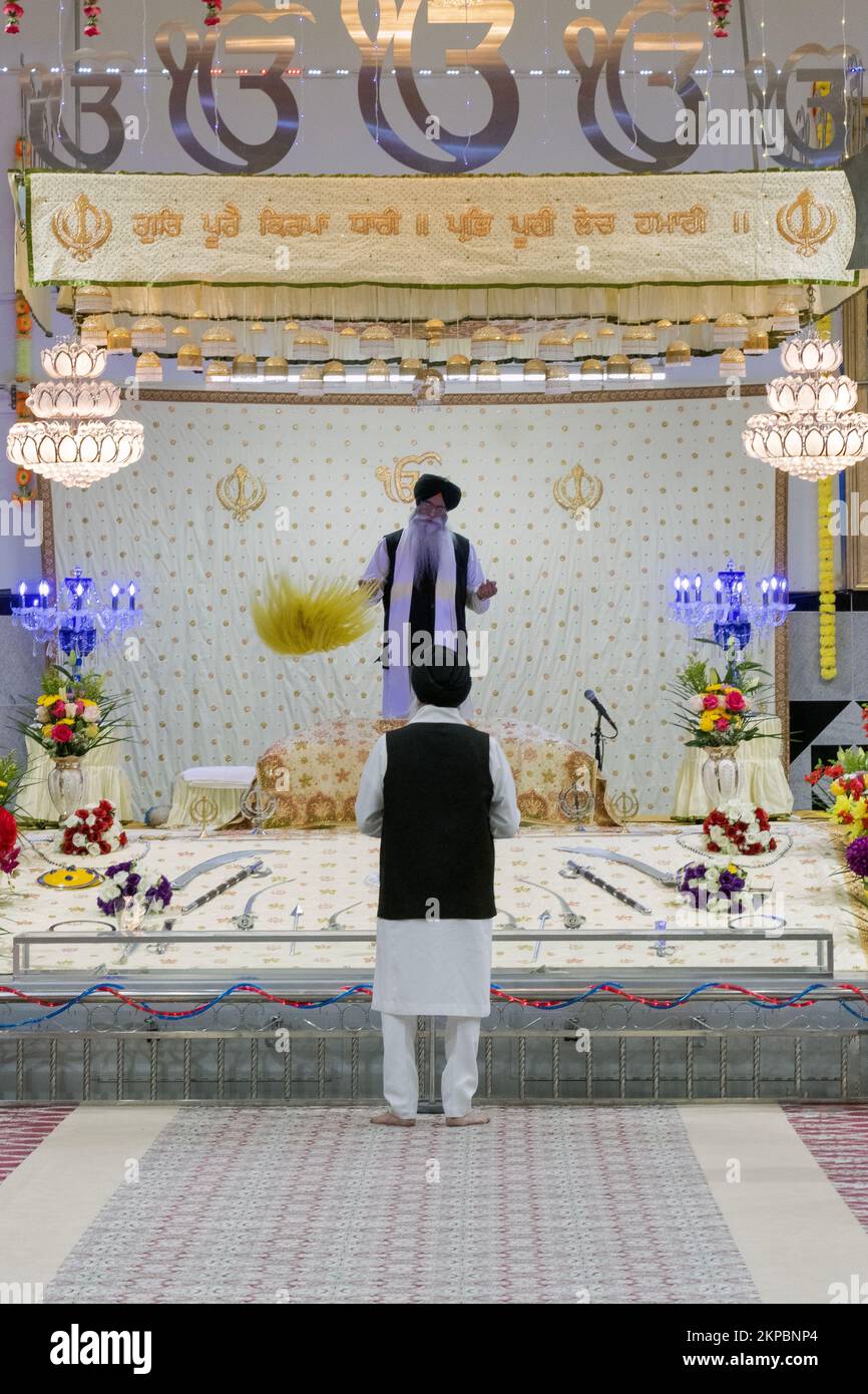 A Sikh pries (foreground) leads a service while a second priest waves his ceremonial yellow flan over the Sikh bible. . At a temple in Queens, NYC Stock Photo