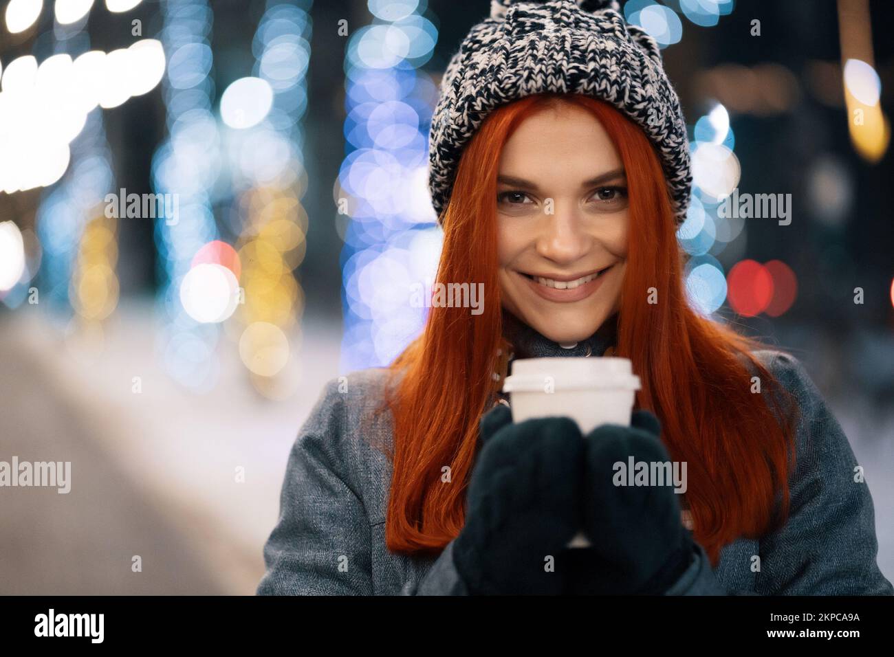 Girl drinking hot coffee while walking in the evening city market decorated with holiday lights in the evening. Feeling happy in big city. Spending wi Stock Photo