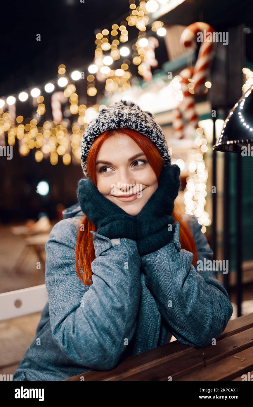 Outdoor night photo of young beautiful happy smiling girl enjoying festive decoration, posing in Christmas fair, in street of european city, wearing k Stock Photo