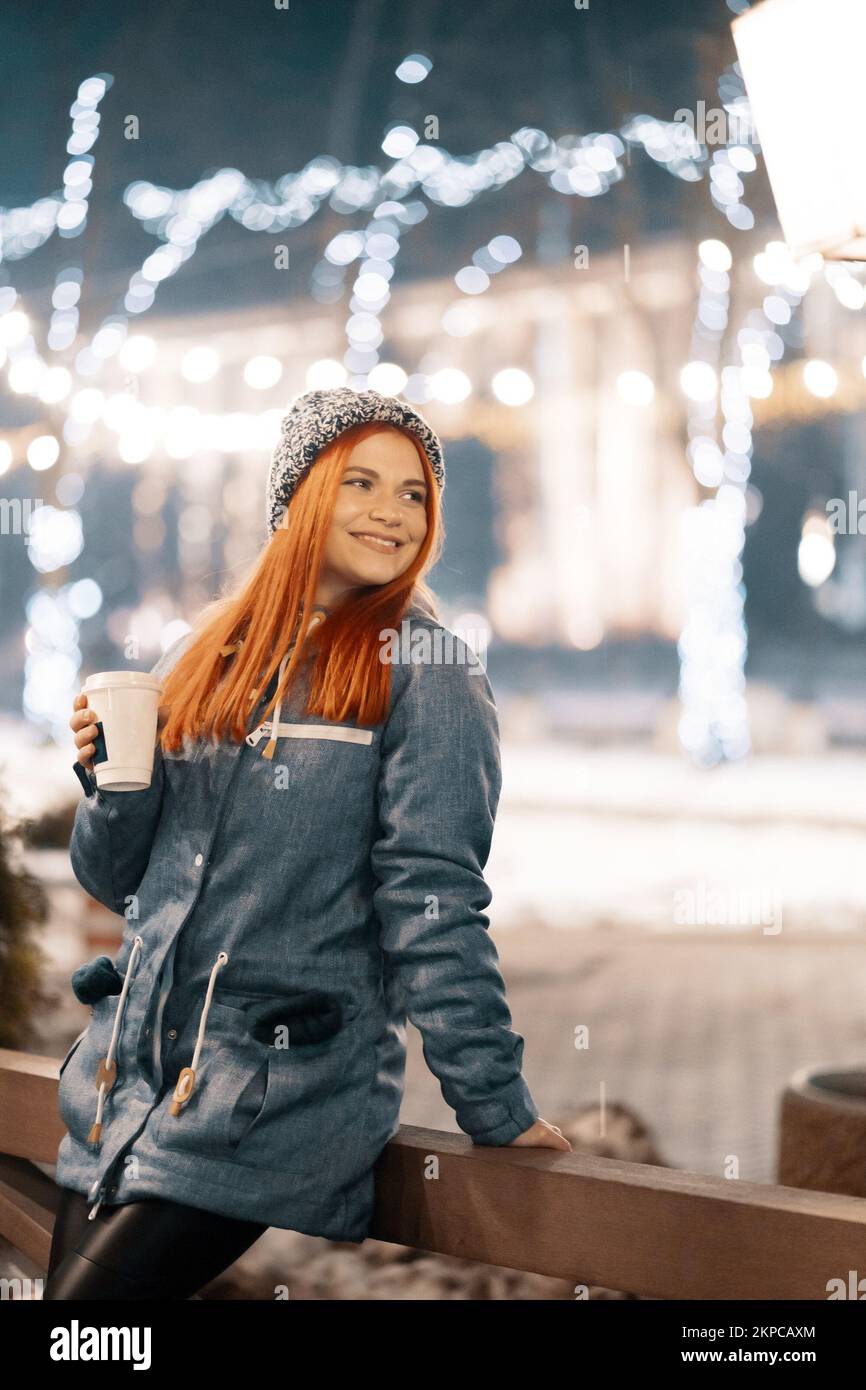 Girl drinking hot coffee while walking in the evening city market decorated with holiday lights in the evening. Feeling happy in big city. Spending wi Stock Photo