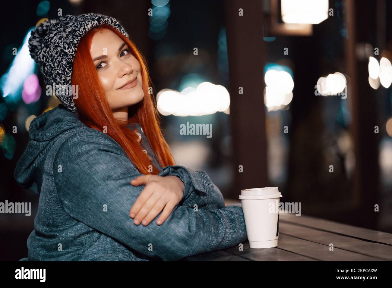 Girl drinking hot coffee while sitting at the table in the evening city market decorated with holiday lights. Feeling happy in big city. Spending wint Stock Photo