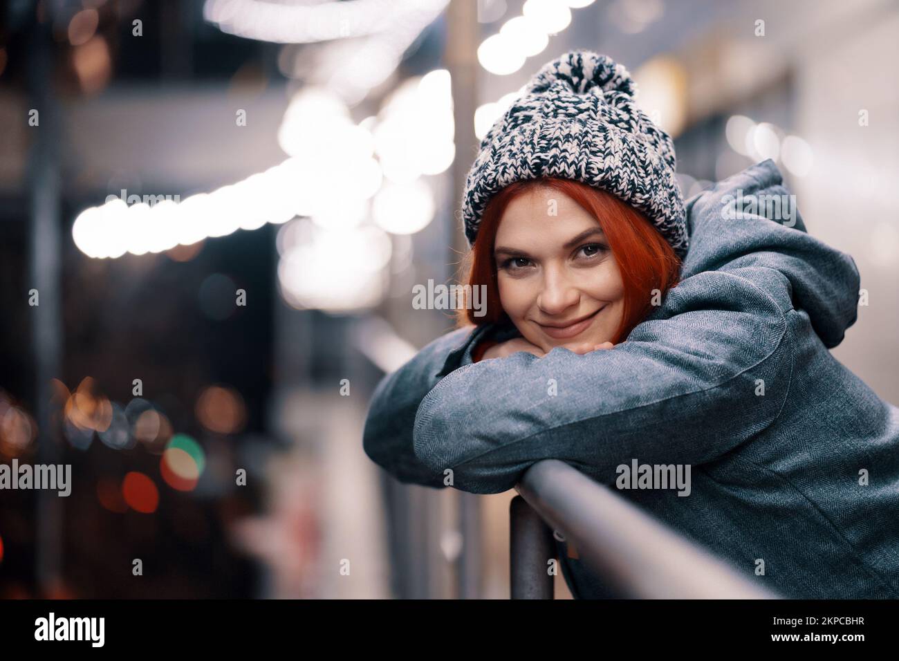 Outdoor night photo of young beautiful happy smiling girl enjoying festive decoration, in street of european city, wearing knitted beanie hat Stock Photo