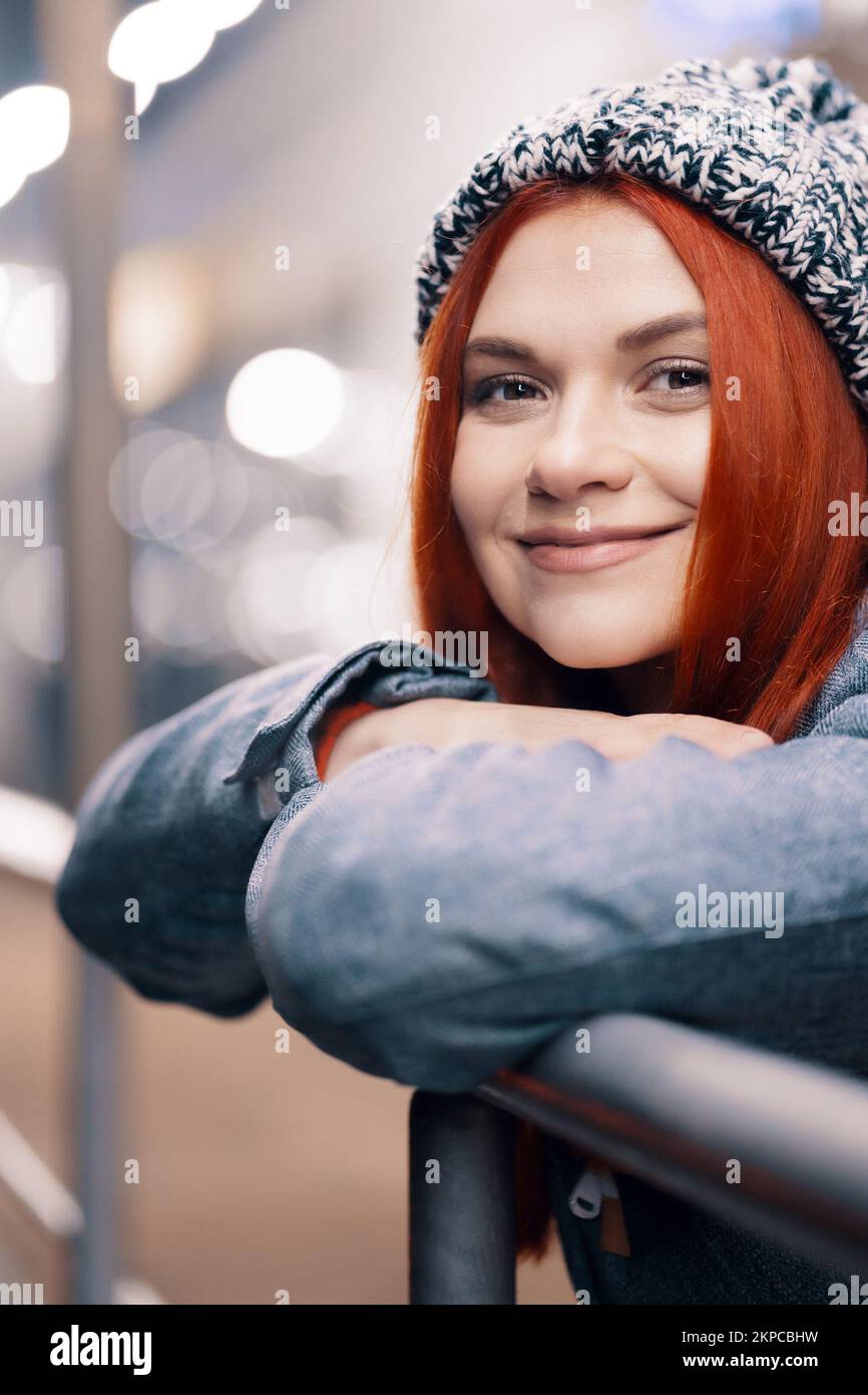 Outdoor night photo of young beautiful happy smiling girl enjoying festive decoration, in street of european city, wearing knitted beanie hat Stock Photo