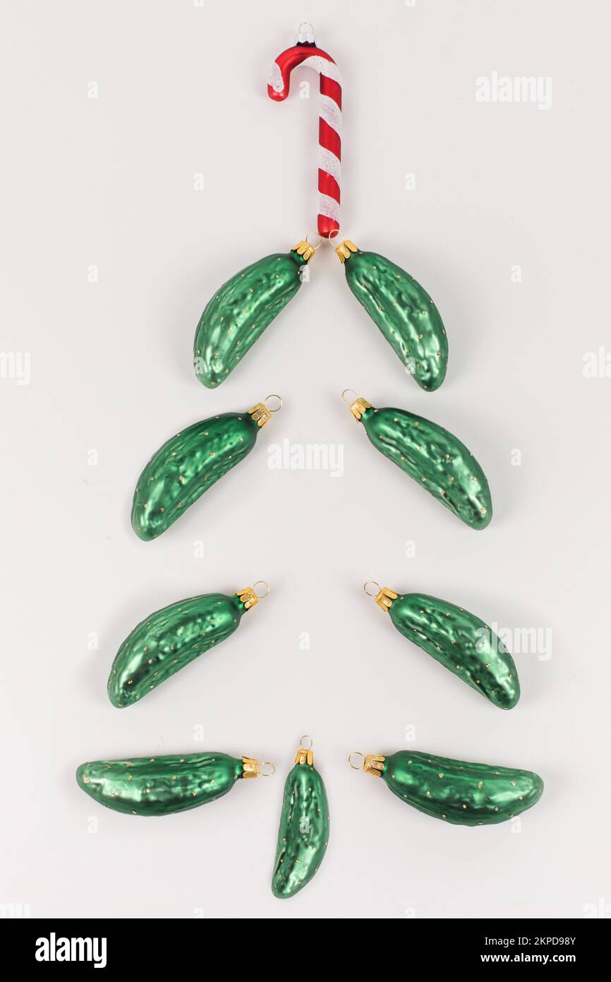 Christmas tree baubles in the shape of a cucumber are lying on a white background. They are arranged like a Christmas tree and a candy cane is on top. Stock Photo
