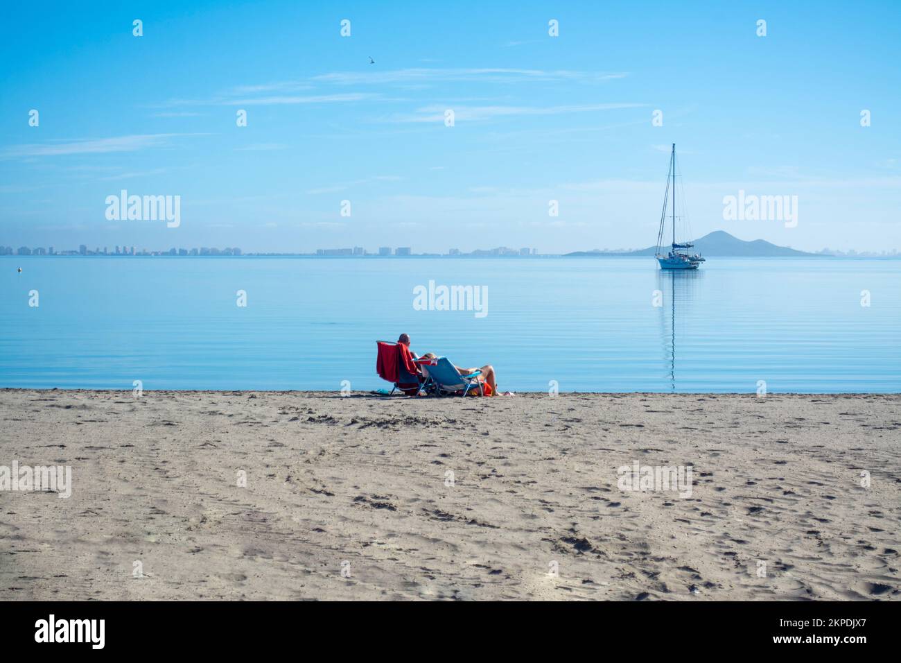 Couple sunbathing on the beach by the calm waters of the Mar Menor with a boat mored out at sea. Stock Photo