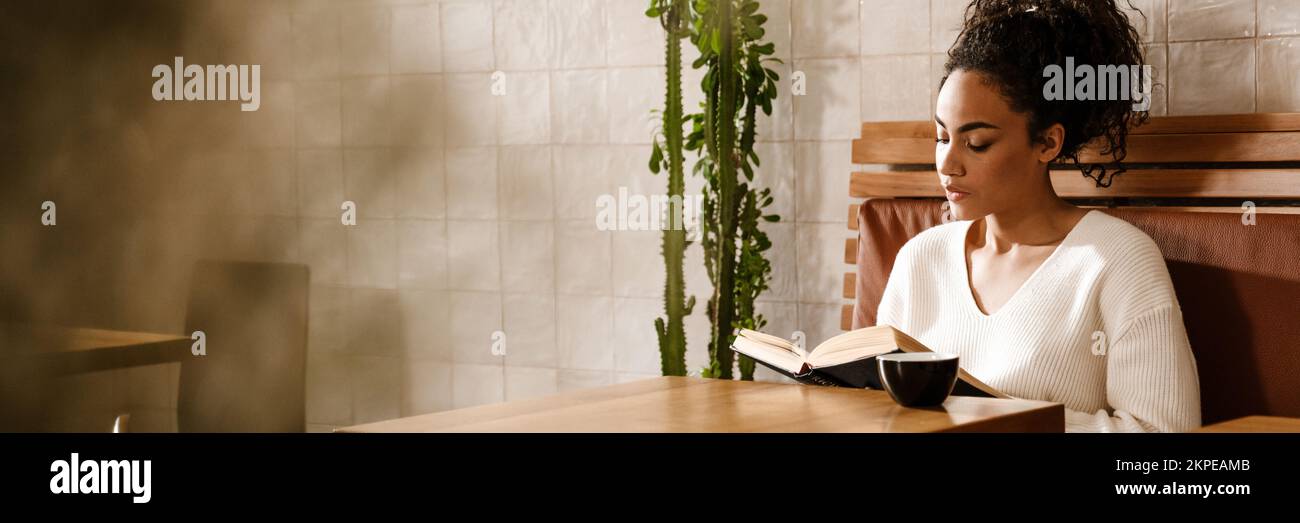 Young black woman drinking coffee and reading book in cafe indoors ...