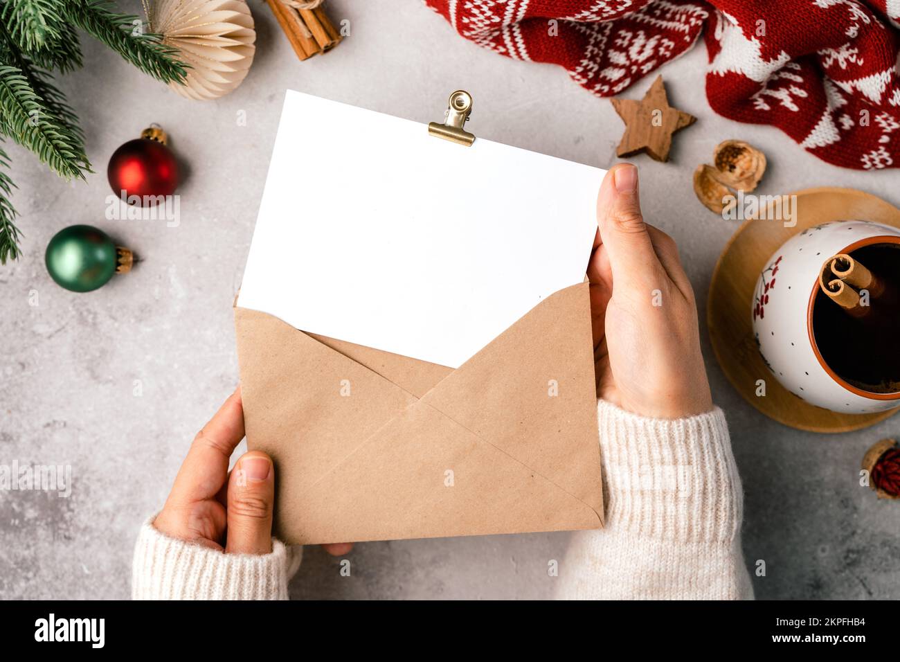 Woman hands in sweater holding blank paper and envelope . Flat lay of gray background with cup of coffee and Christmas decoration. Top view mock up Stock Photo