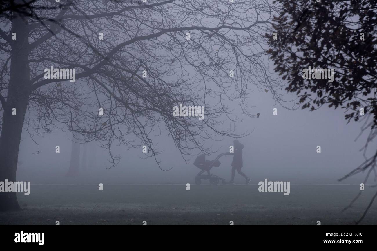 Harrogate, UK. 28th Nov, 2022. An eerie landscape was created by the fog on the Stray in the town centre this morning. A lady with a pram is silhoutted in the strange light. Picture Credit: ernesto rogata/Alamy Live News Stock Photo